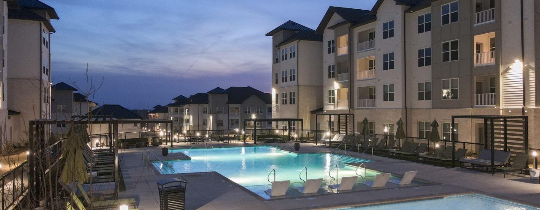 Luxury resort-style swimming pool at The Ravelle at Ridgeview apartments near Nashville, featuring modern poolside lounge chairs, private cabanas, and upscale apartment buildings in the background. The pool is illuminated at dusk, highlighting the high-en
